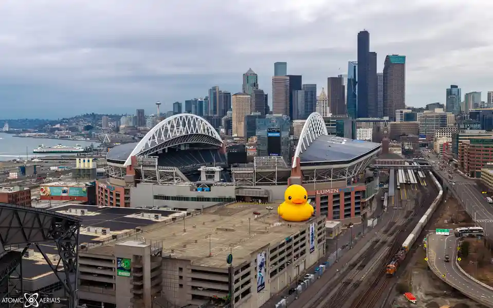 An aerial photo of Seattle's Lumen Field stadium with a giant yellow rubber duck placed on top of a parking garage in front…