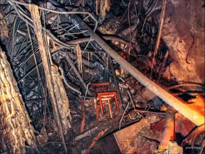 a red plastic chair sits alone in the middle of a dark, tangled mess of burnt debris and twisted metal, looking out of place…