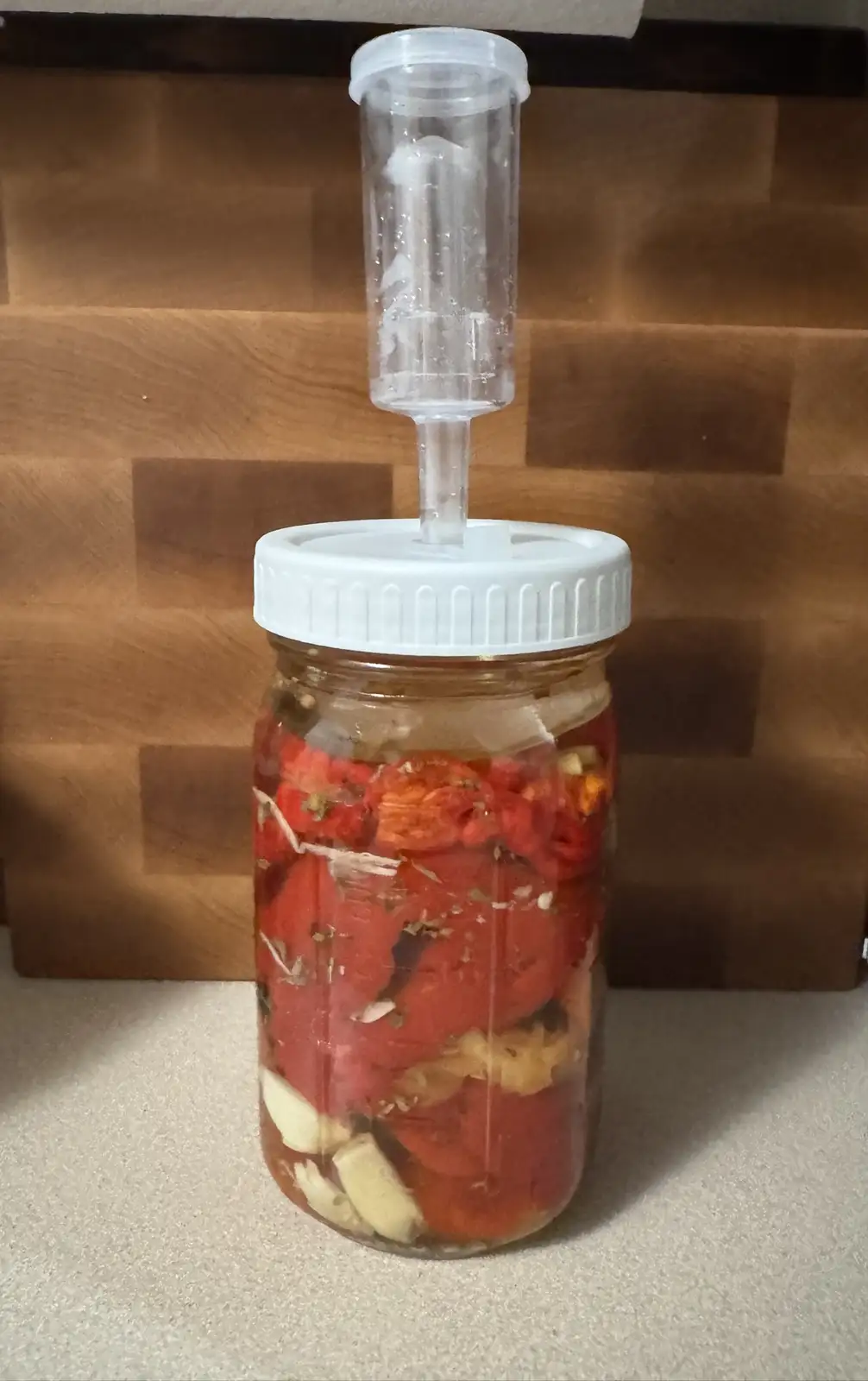 A glass mason jar filled with chopped red peppers, garlic cloves, and herbs submerged in liquid, sealed with a white lid and…