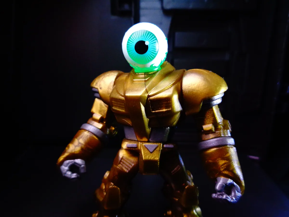 A golden, armored action figure stands in a dark setting, its head replaced by a large, glowing green eyeball with a black…
