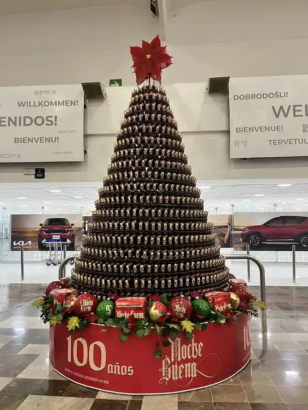 A large Christmas tree made entirely of stacked Morche Brem beer bottles stands in an airport terminal, topped with a red…