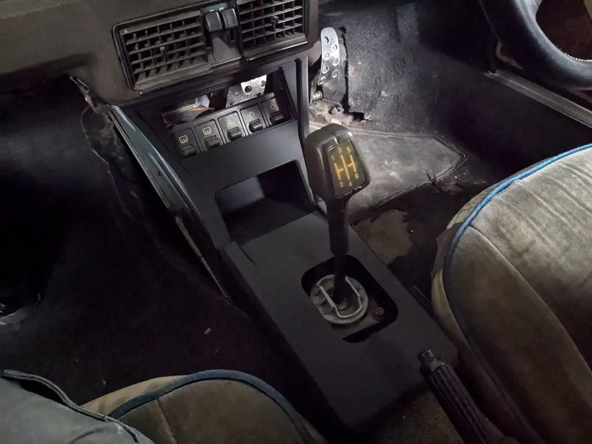 A close-up shot of a car's interior focusing on a manual gear shifter with a lit H-pattern display, surrounded by worn…