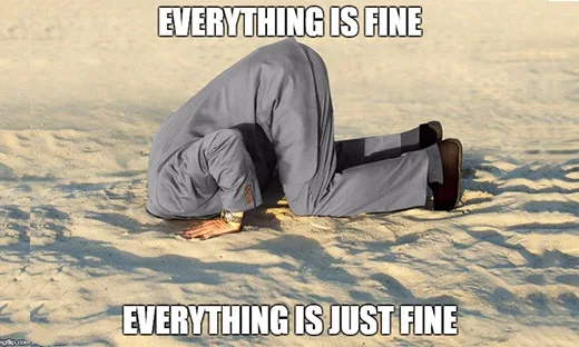 A person in a grey suit is kneeling on a sandy beach with their head buried in the sand, conveying a sense of hiding or…