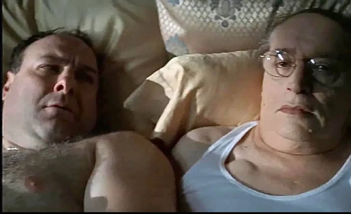 two men lie in bed, one shirtless with a stern expression, the other in a white tank top with glasses and a smug look, both…
