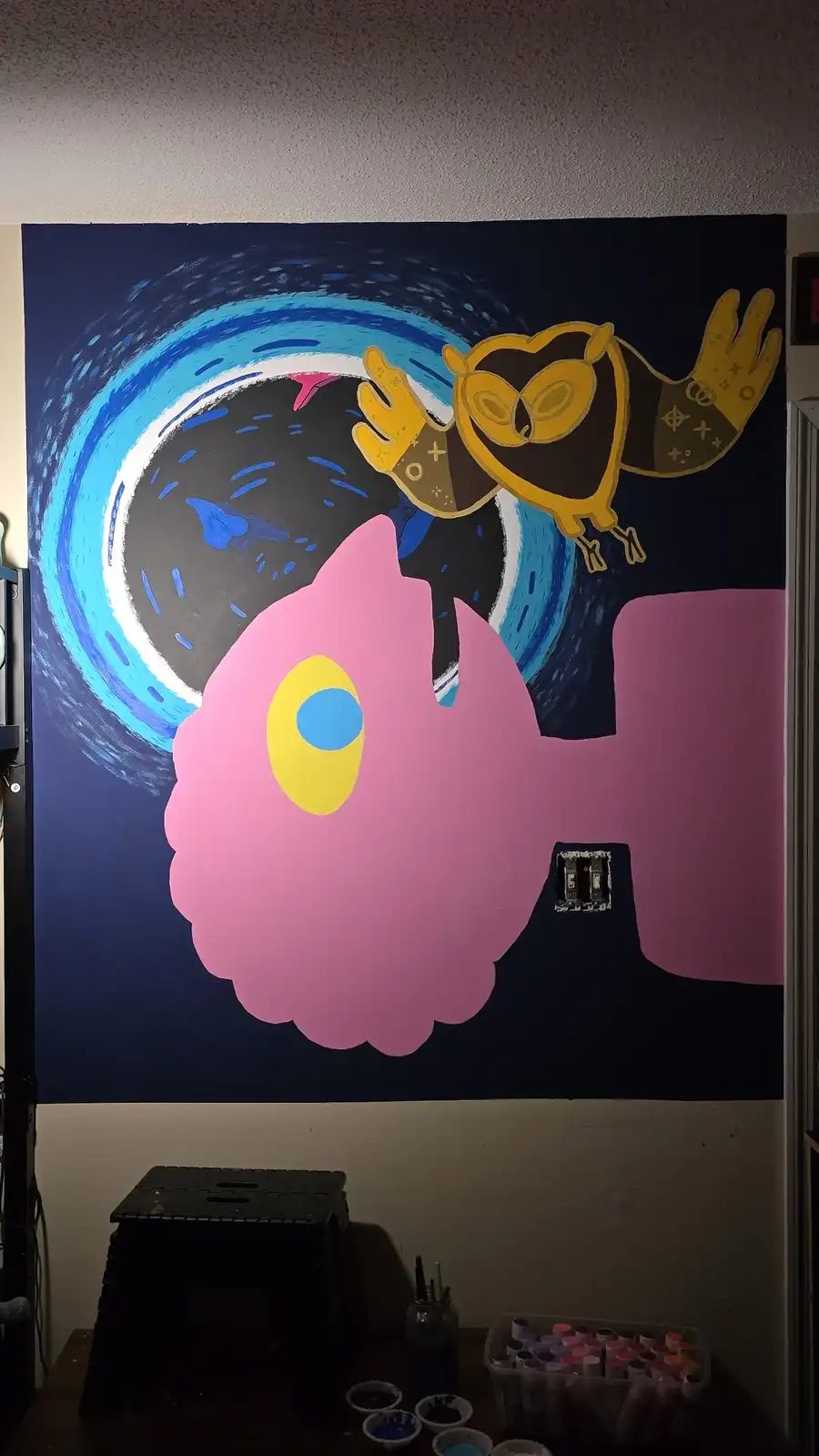 A hand-painted wall mural depicts a stylized pink creature with a large yellow eye and a yellow owl-like figure with…