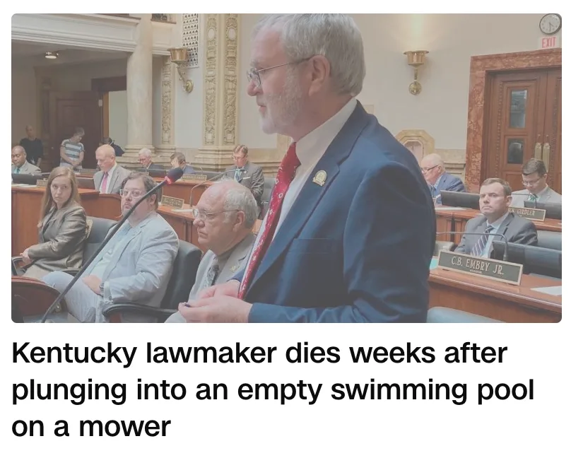 A man in a suit and tie stands at a podium in a formal legislative chamber, speaking into a microphone while other officials…