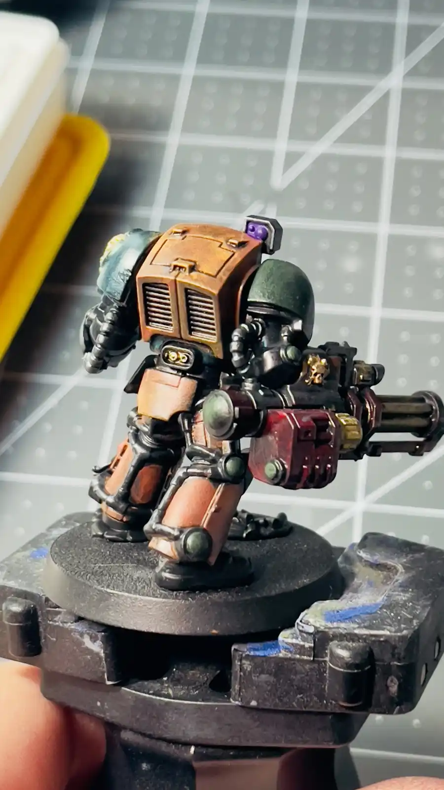A close-up photo shows a hand holding a painted miniature of a heavily armored, futuristic soldier with a large weapon…