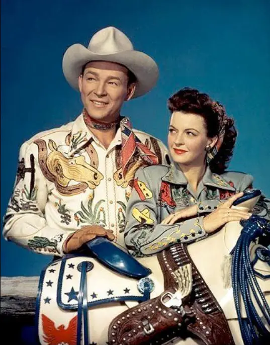 A vintage promotional photo of a smiling man and woman in ornate cowboy and cowgirl attire, posed with a saddle and rope…