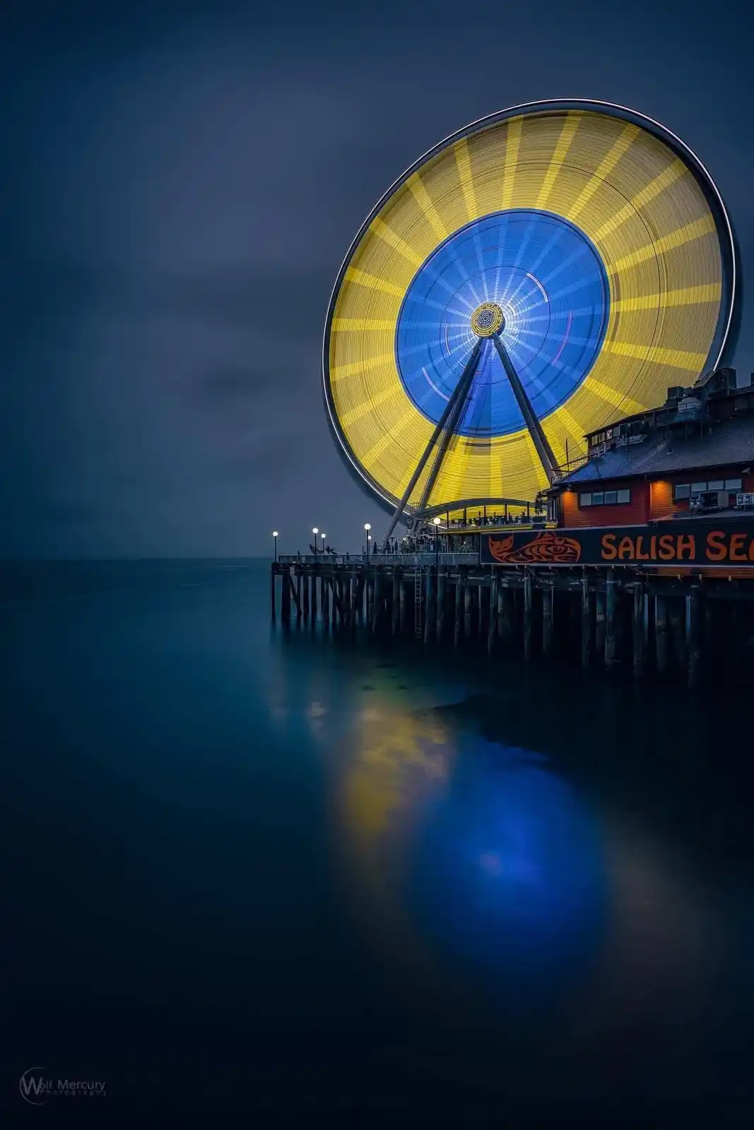 A nighttime photograph of the Seattle Great Wheel, a large illuminated Ferris wheel with yellow and blue lights, standing on…