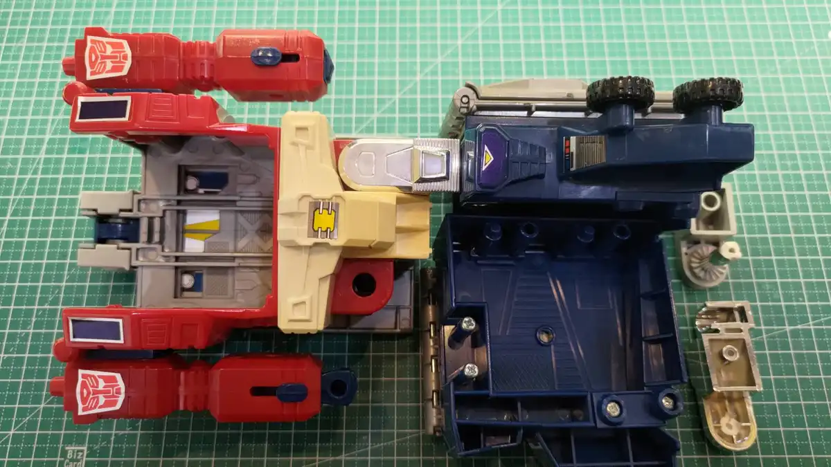 Two disassembled toy robots, one red and one blue, lie on a green cutting mat with their internal mechanisms exposed. The…