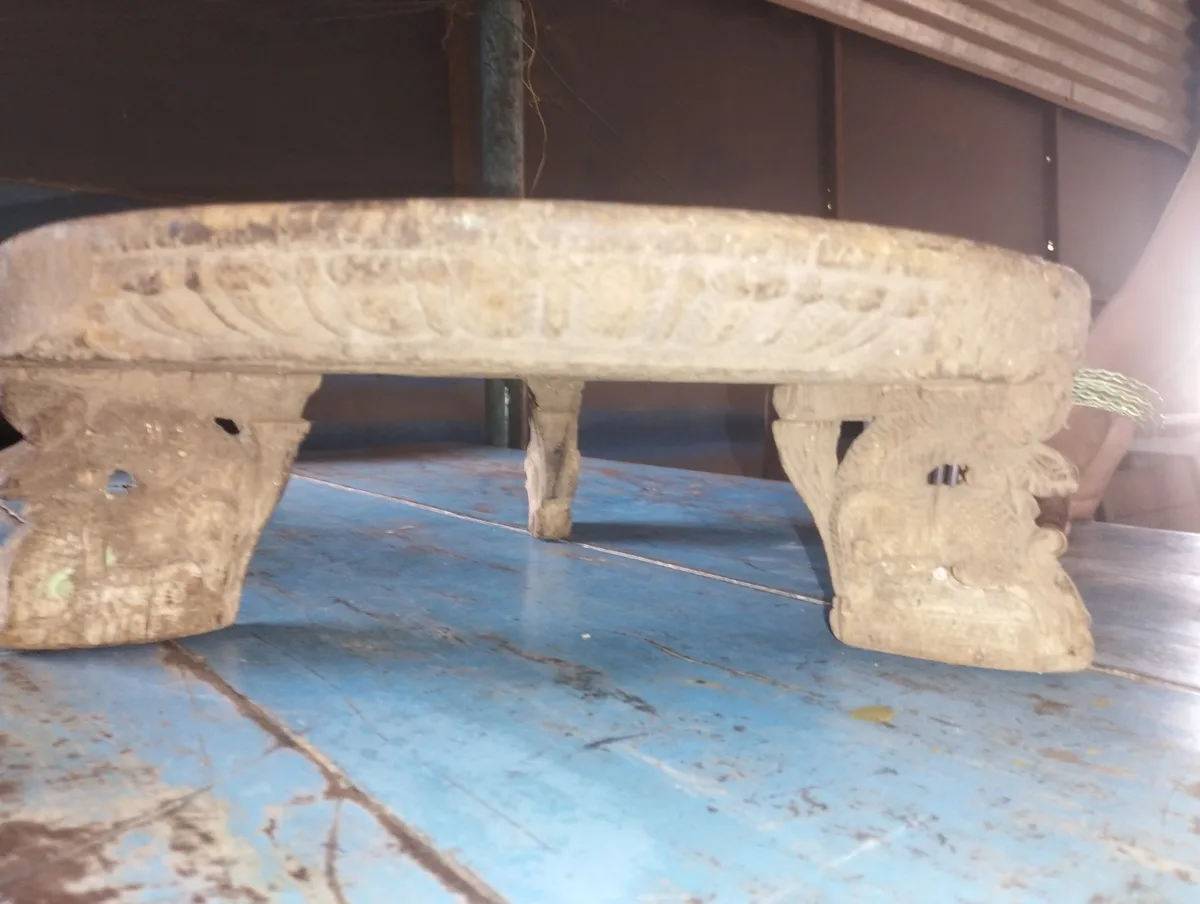 A low, ornate wooden stool with carved legs sits on a worn blue wooden floor. The stool appears old and weathered, with…