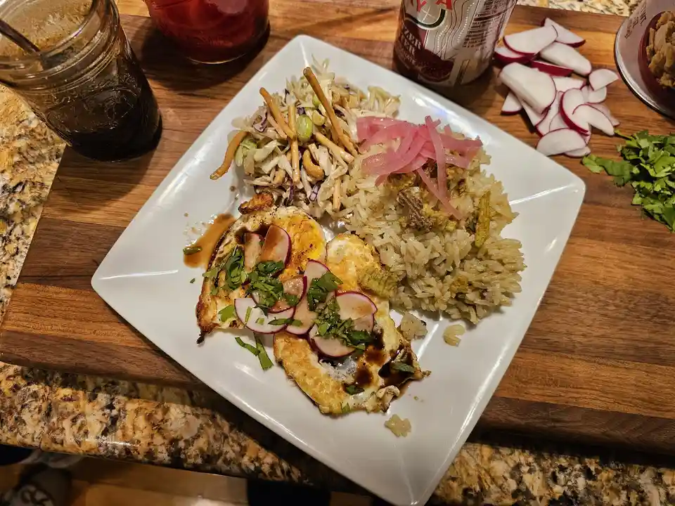 A white square plate holds a meal of fried eggs topped with sliced radishes and cilantro, served with rice and a…