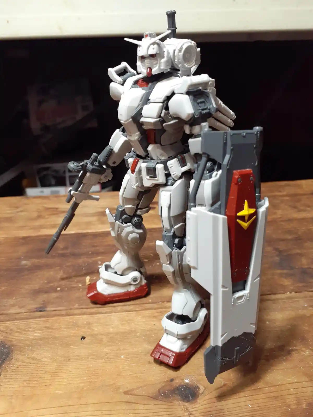 A white, grey, and red Gundam model stands on a wooden table holding a rifle and a shield with a yellow star. The overlaid…