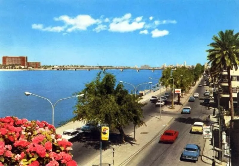 A vintage postcard-style photo of a sunny city street beside a wide river, with palm trees, classic cars, and a distant…
