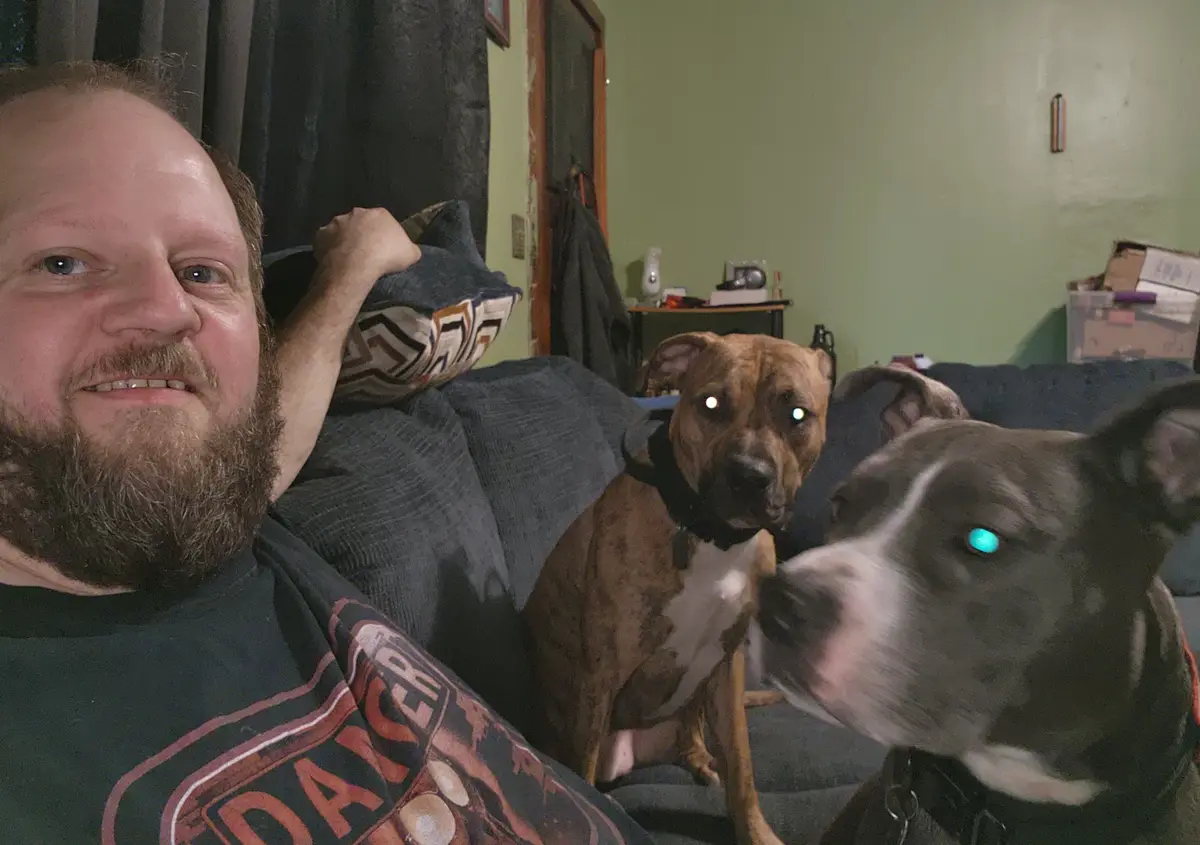 A man with a beard smiles at the camera while sitting on a couch with two dogs. One dog has normal glowing eyes, while the…
