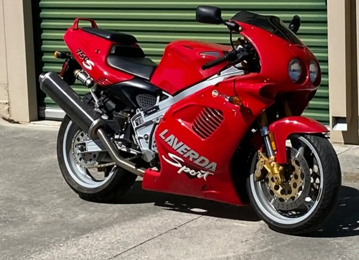 A red Laverda Sport 750S motorcycle is parked outdoors in front of a green roll-up door. The bike has a sporty design with…