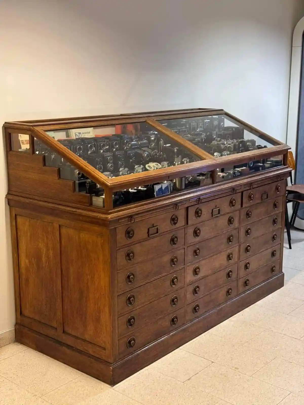 A large, antique wooden display cabinet with a glass top and many small drawers below, filled with numerous vintage cameras…