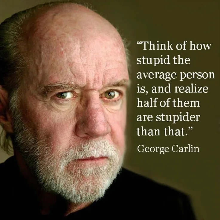 A close-up portrait of George Carlin, an older man with a bald head, white beard, and intense, slightly narrowed eyes…