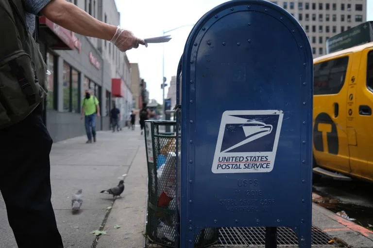 A person wearing a plastic glove holds a knife over a blue United States Postal Service mailbox on a city sidewalk. A pigeon…