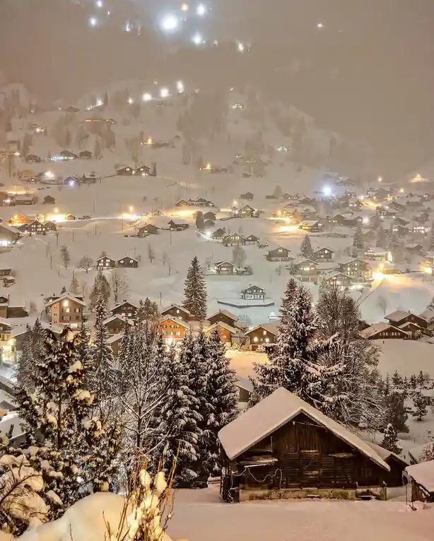 A snowy, nighttime landscape of a mountain village with illuminated wooden chalets and ski resort lights on the slopes. The…