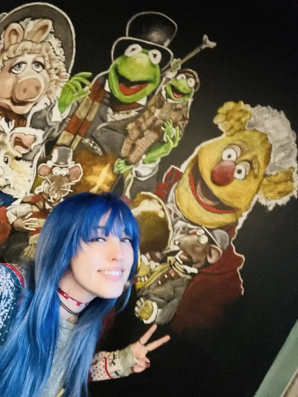 A person with vibrant blue hair and bangs smiles at the camera while posing in front of a large, colorful mural depicting…