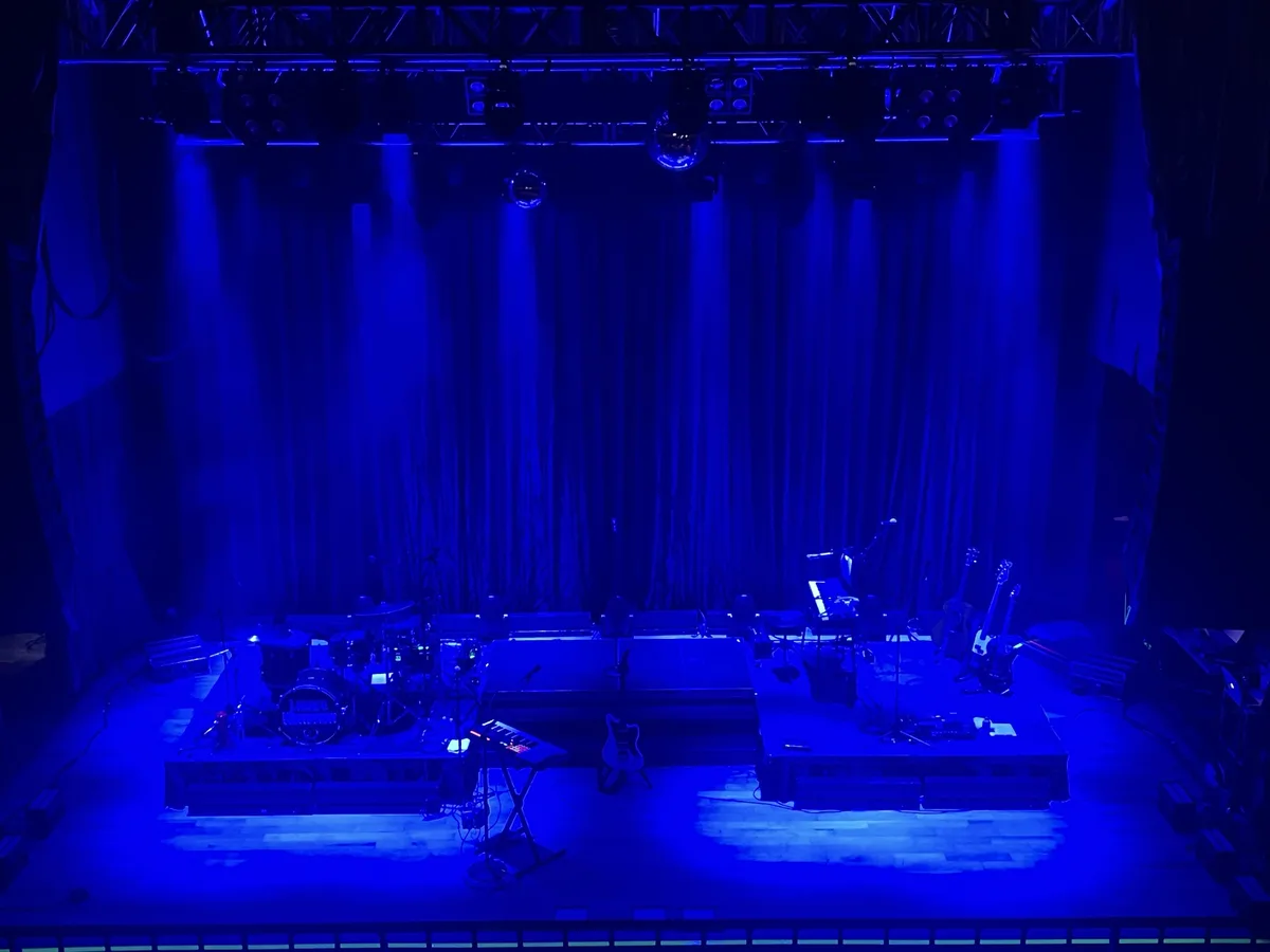 A dimly lit concert stage bathed in blue light, featuring a drum set, keyboards, and guitars arranged on platforms, with…