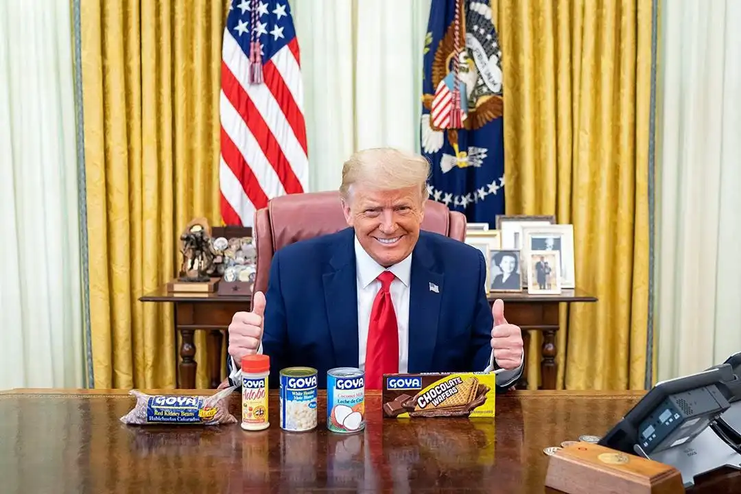 A man in a suit and red tie sits at a desk in an official-looking room with American flags behind him, giving two thumbs up…