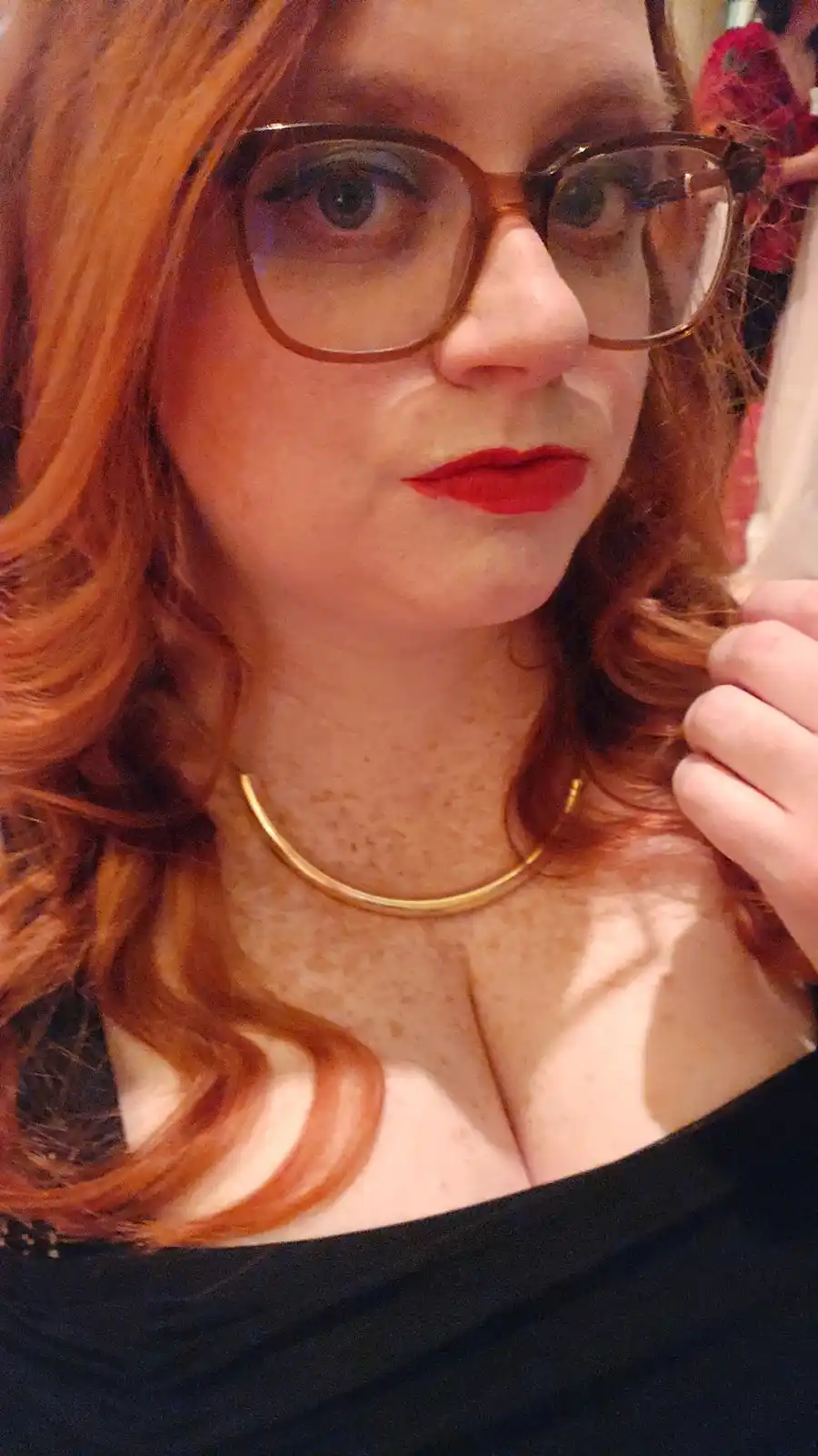 A close-up selfie of a woman with red hair, wearing large brown-framed glasses, bright red lipstick, and a gold choker…