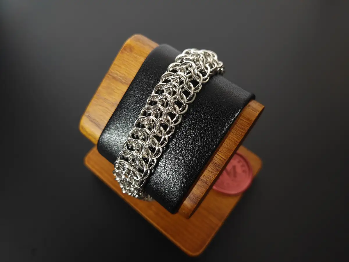 A silver chainmaille bracelet is displayed on a black leather-wrapped wooden stand against a dark background. The bracelet…