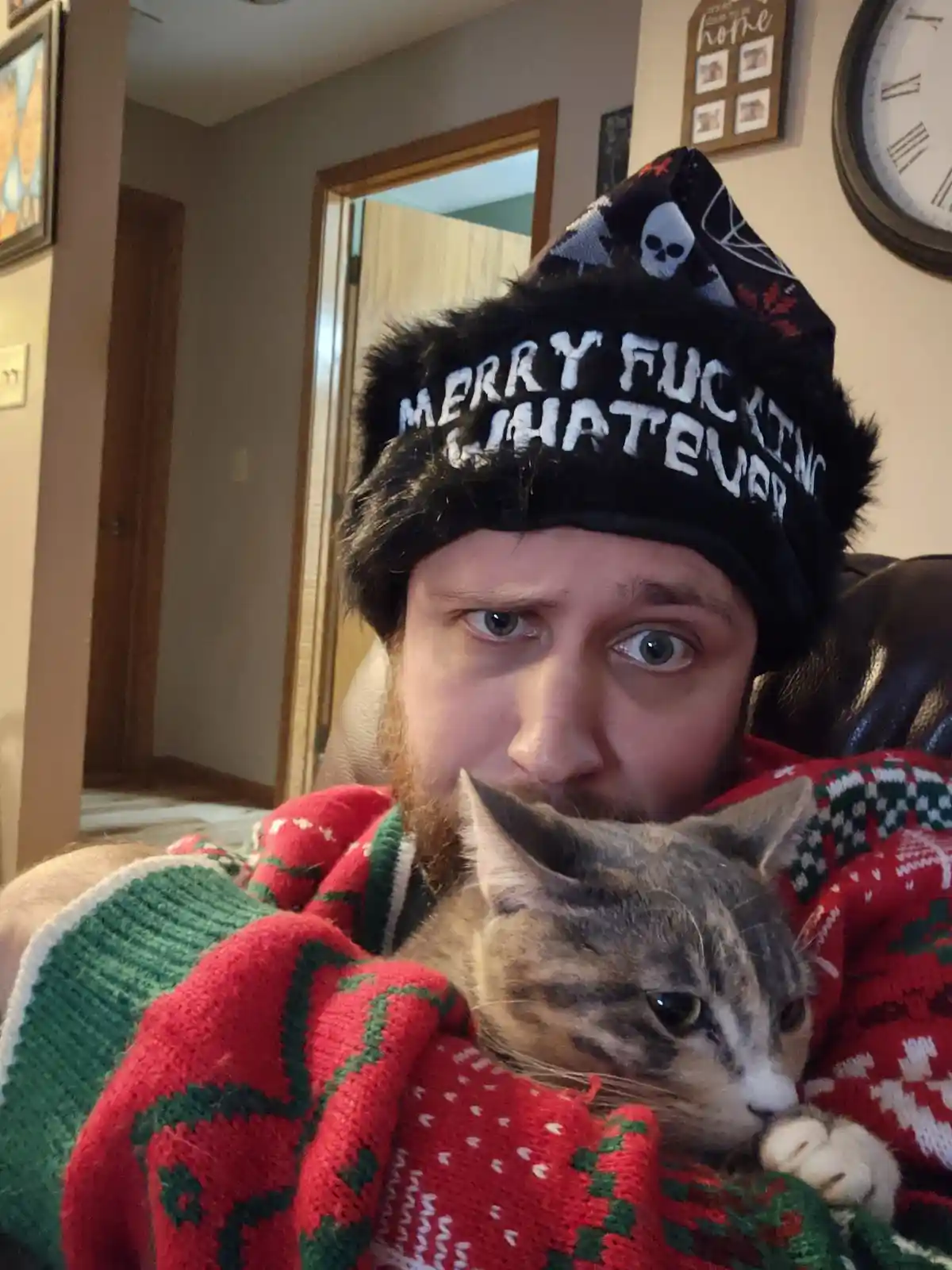 A man with a beard wears a black, furry winter hat with the text 'MERRY FUCKING WHATEVER' and a skull design, while wrapped…