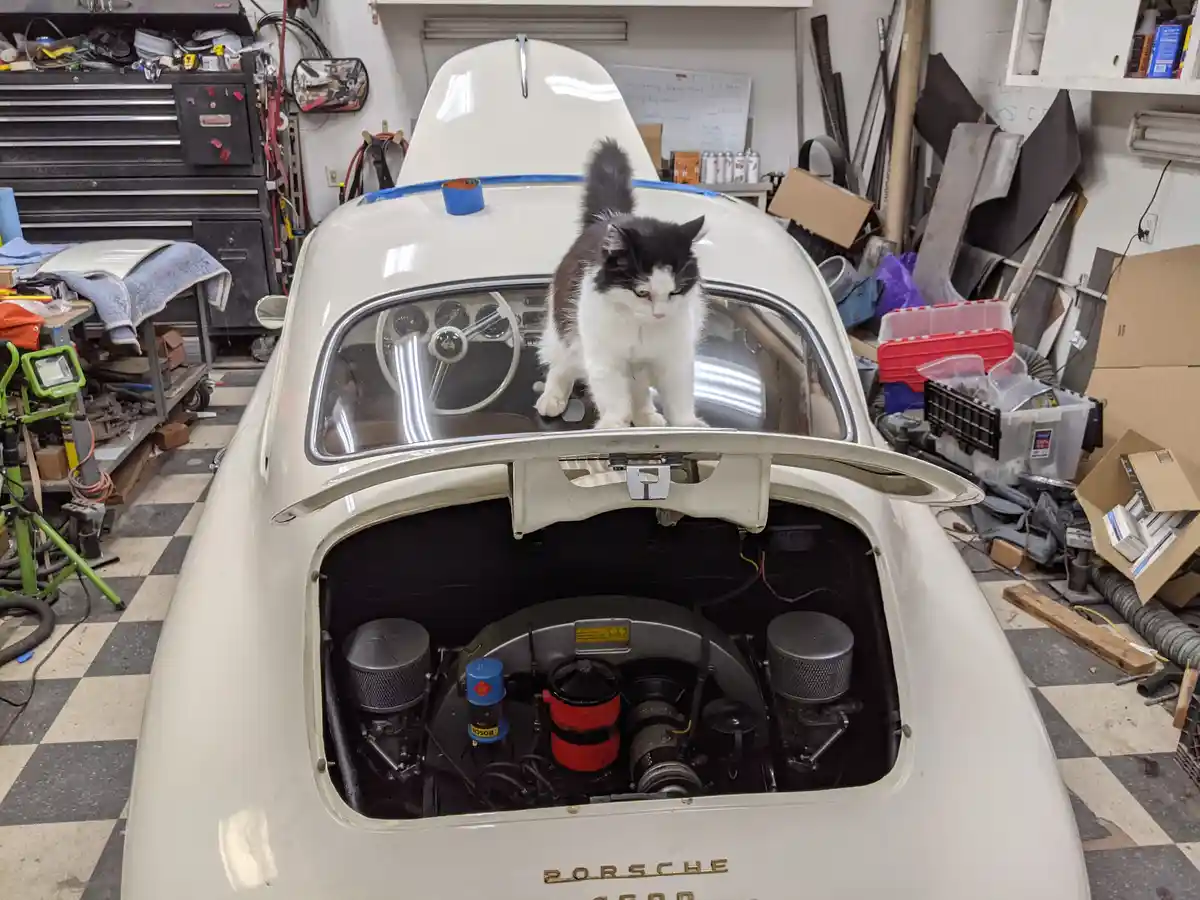 A black-and-white cat stands atop the open hood of a white vintage Porsche 356, looking down at the engine. The car is in a…