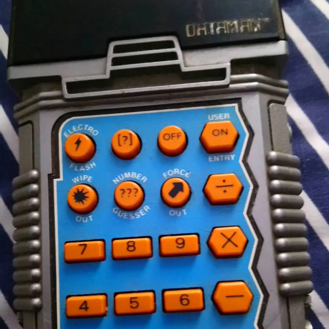 A close-up of a vintage Dataman handheld electronic device with a blue keypad featuring orange buttons labeled with…