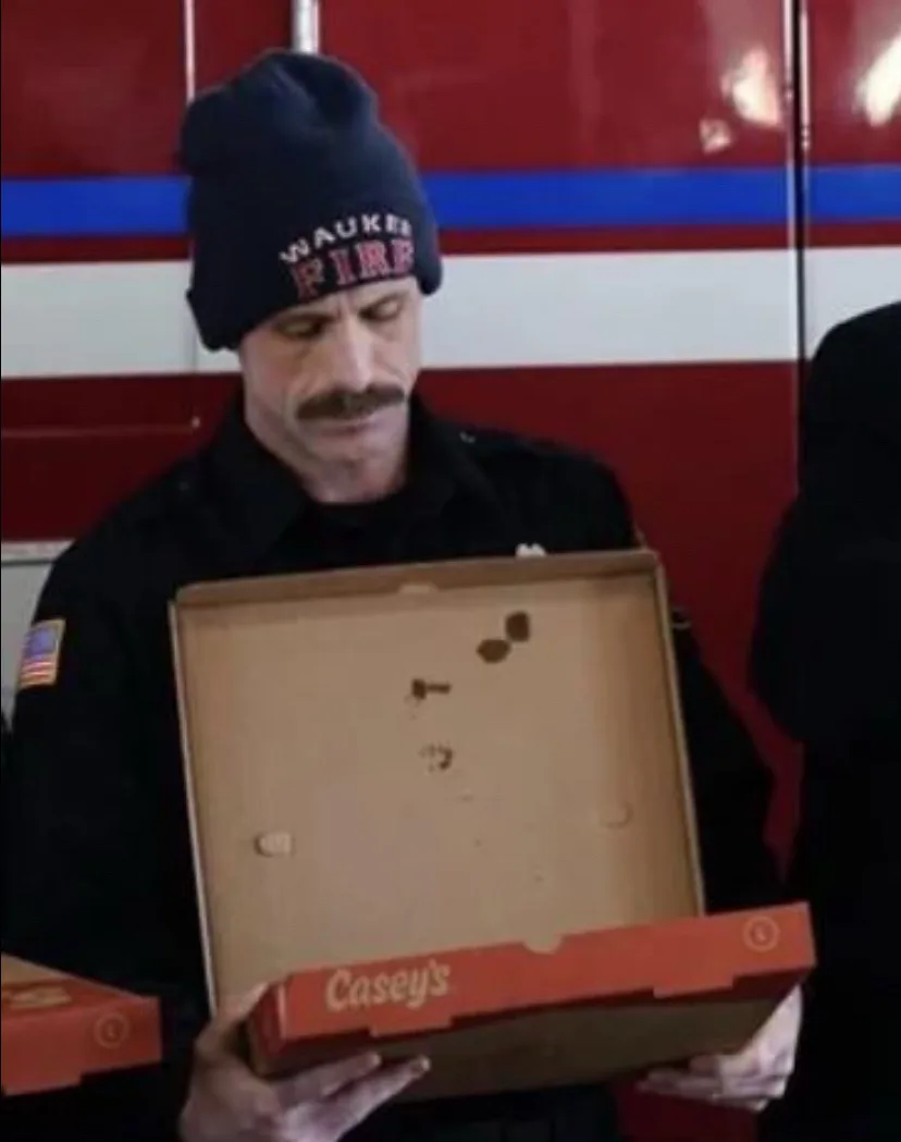 A man in a firefighter uniform and a beanie holding an empty pizza box from Casey's, looking down at it with a confused or…
