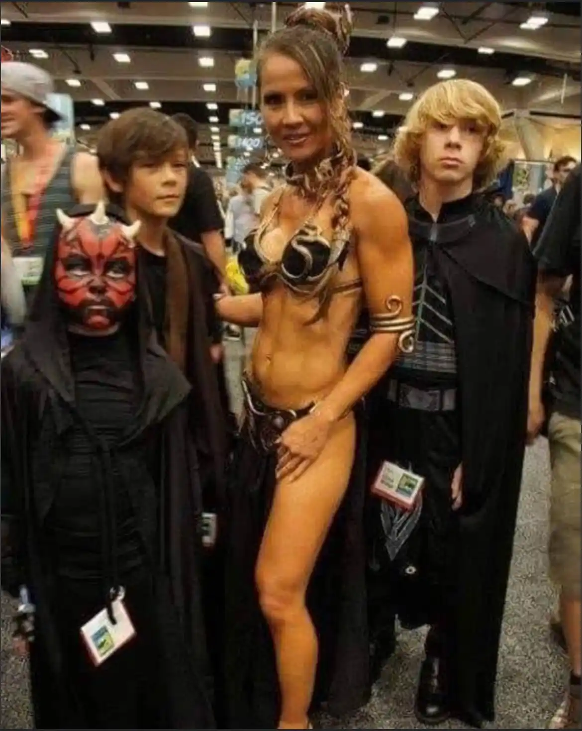 Three people in Star Wars cosplay pose for a photo at a convention: a child in a Darth Maul mask and robe, a woman in a…