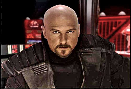 A bald man with a goatee, wearing a dark armored jacket, stares intensely at the viewer. Behind him, red-lit control panels…