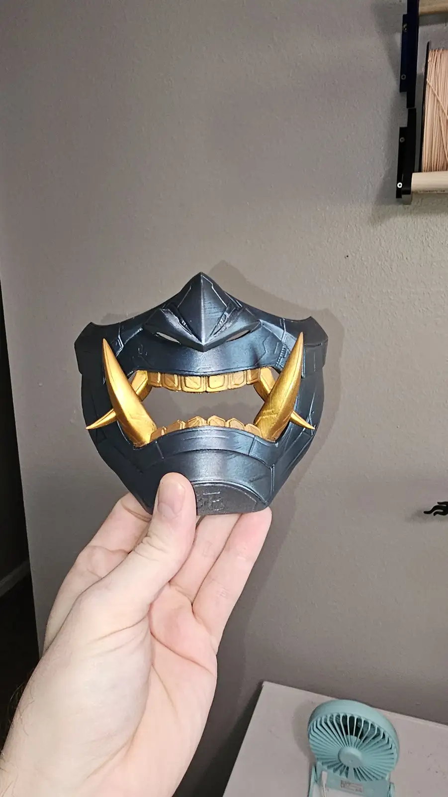 A hand holds up a custom-made, dark gray and gold mask with sharp, pointed teeth and horns, resembling a fantasy or anime…