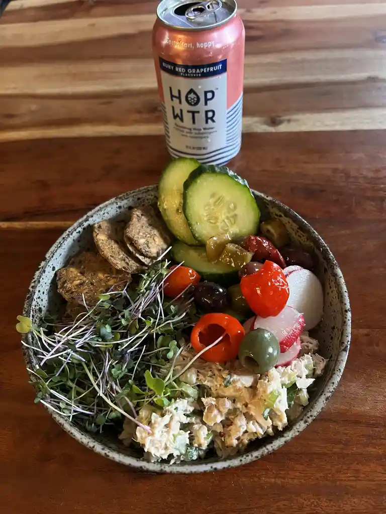 A speckled ceramic bowl filled with a colorful salad of tuna, microgreens, cucumber slices, cherry tomatoes, olives…