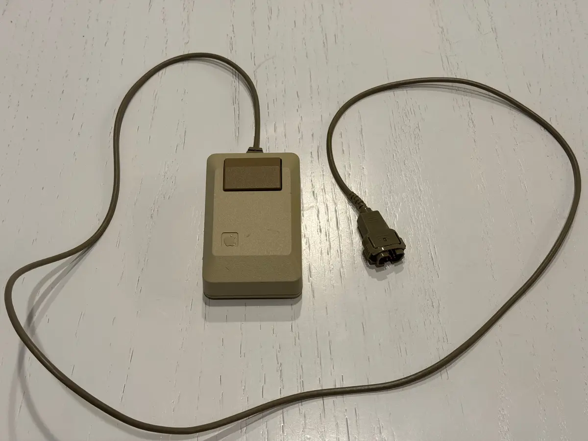 A beige, rectangular Apple mouse from the 1980s with a single button and a thick cord lies on a white wooden surface. The…