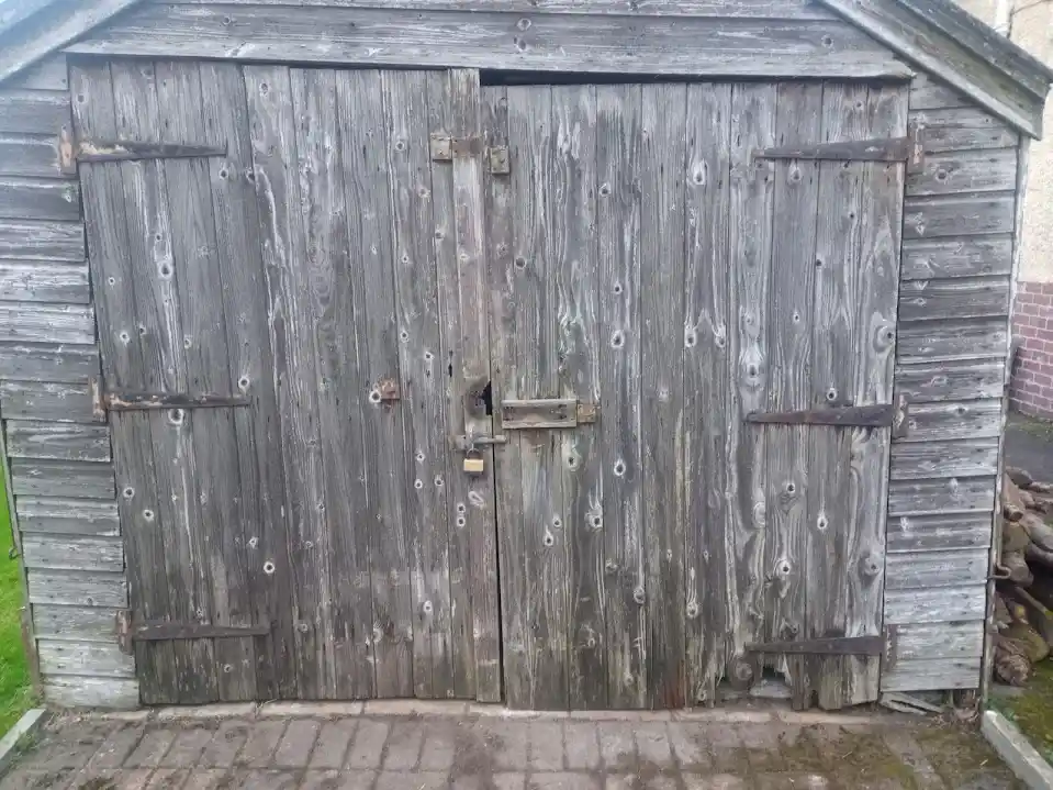 A weathered wooden shed with double doors, one slightly ajar, showing a rusty lock and multiple holes in the wood. The…