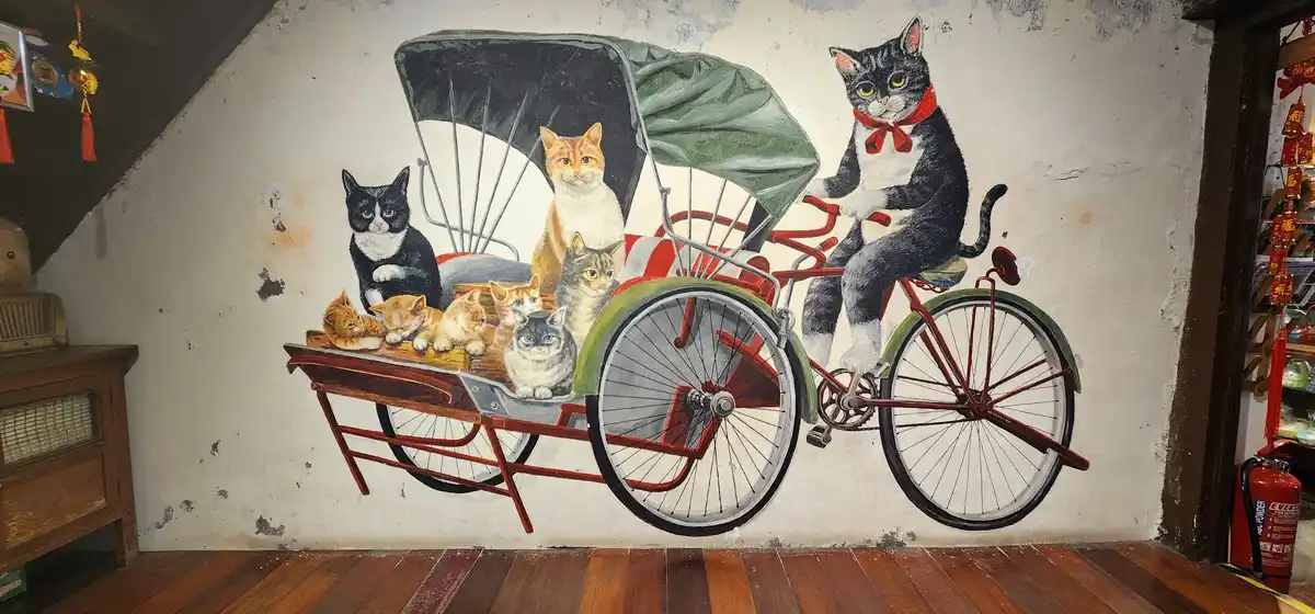 A mural on a wall depicts a black-and-white cat wearing a red bow tie, pedaling a red tricycle rickshaw with a green canopy…