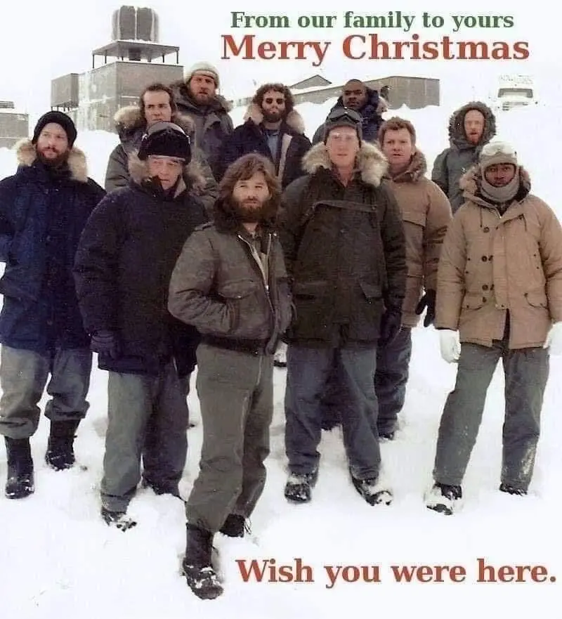 A group of men in heavy winter coats and hats stand in a snowy outdoor setting, possibly a remote station, with buildings…