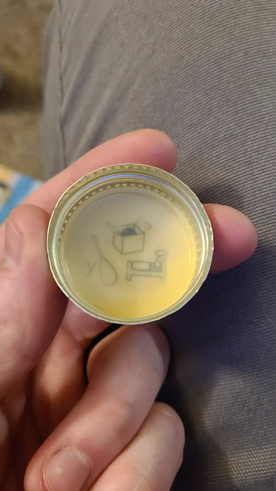 A hand holds a bottle cap with a faded, etched design showing a box with a dark object inside, a spoon, and a bed with a…