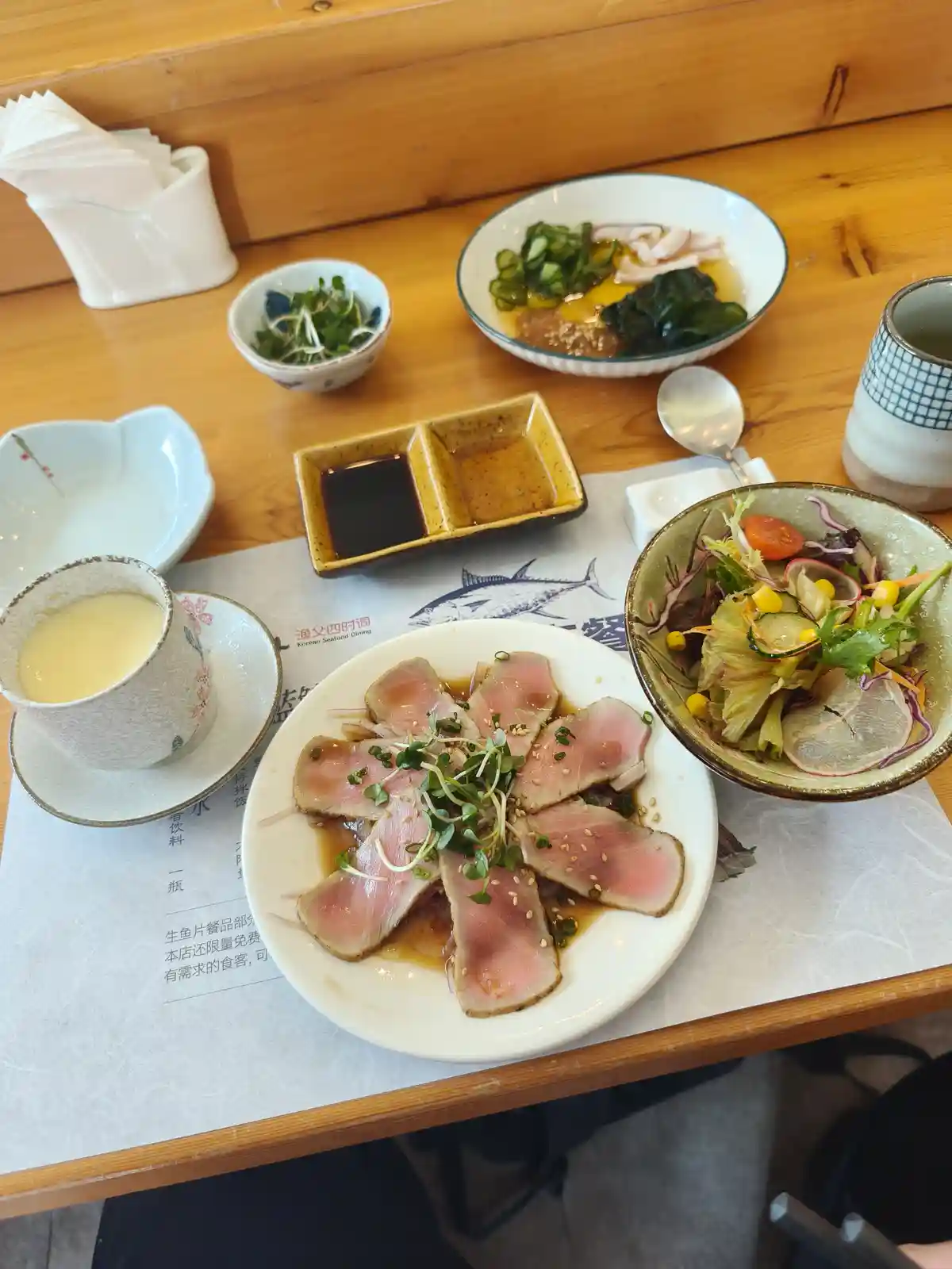 A table at a restaurant is set with a full meal including a central plate of seared tuna sashimi garnished with microgreens…