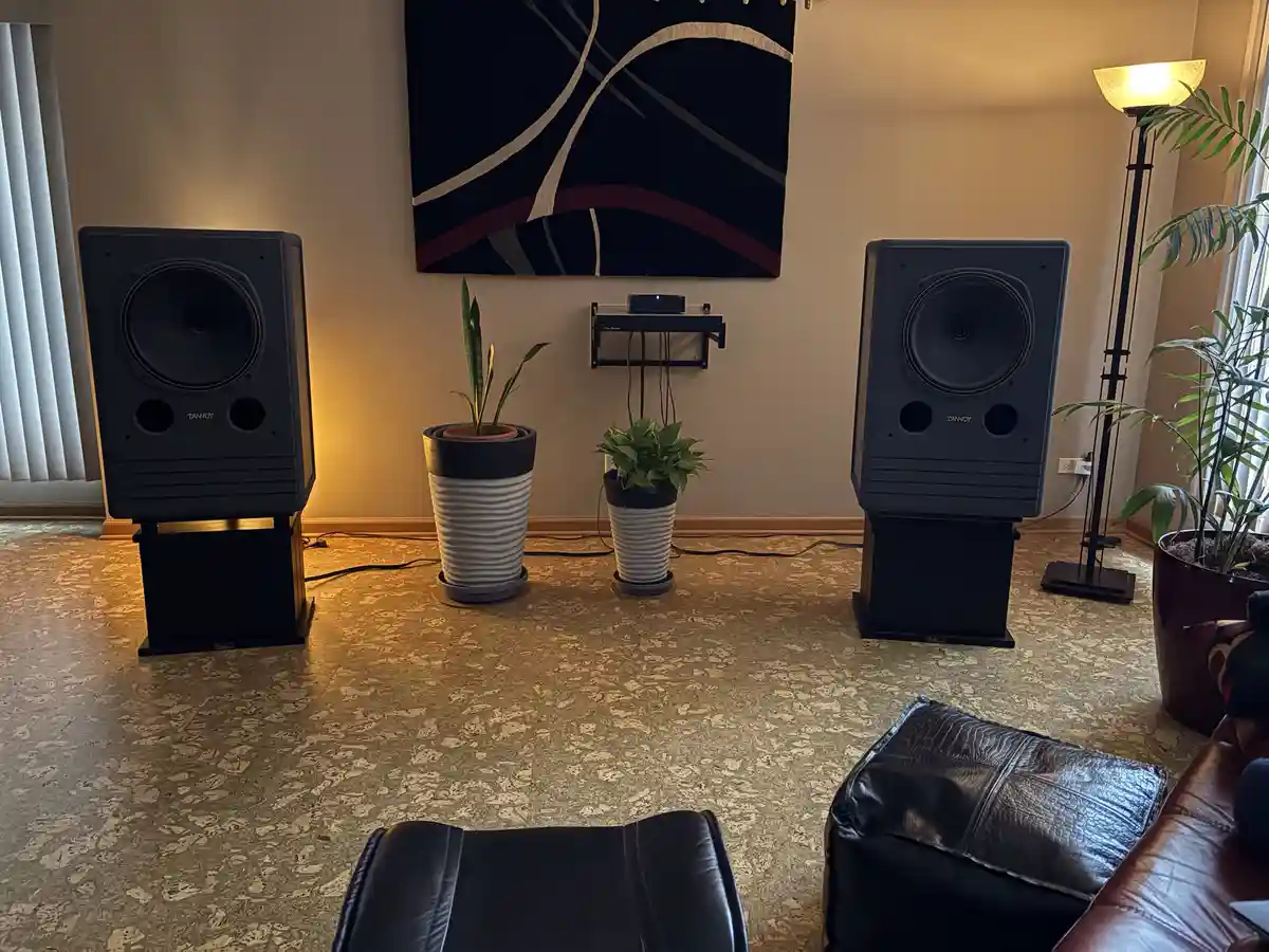 A cozy living room features two large, black Tannoy floor-standing speakers flanking a small shelf with a soundbar, set…