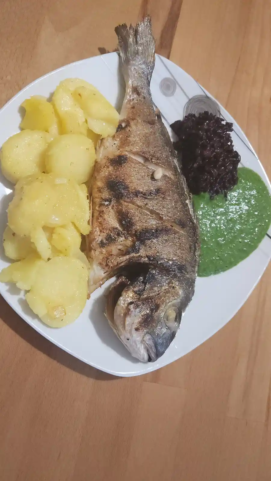A whole grilled fish is served on a white plate alongside boiled potatoes, black rice, and a green sauce. The fish has…