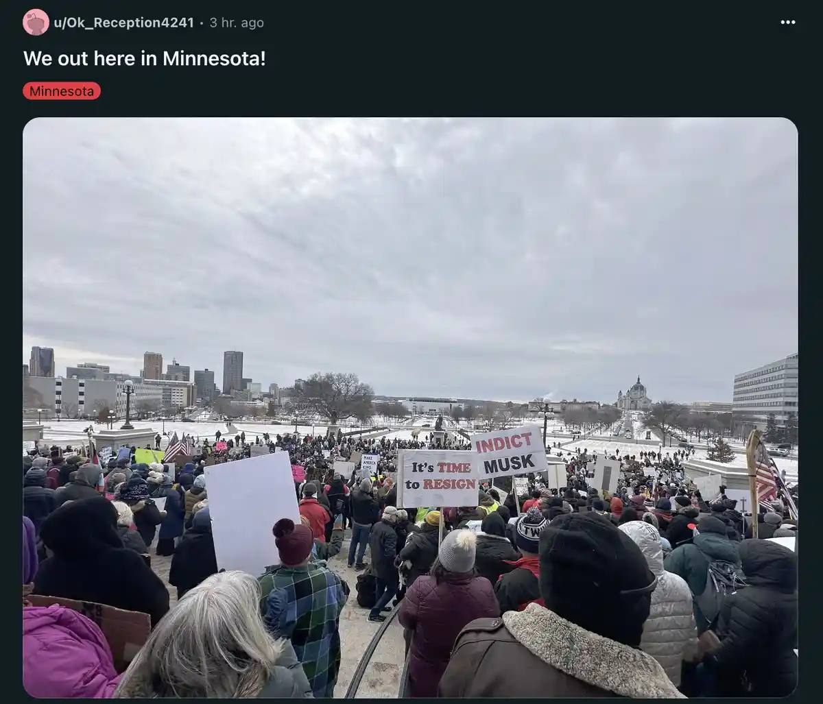 A large crowd of people dressed in winter clothing gathers outdoors in a snowy urban plaza, holding protest signs including…