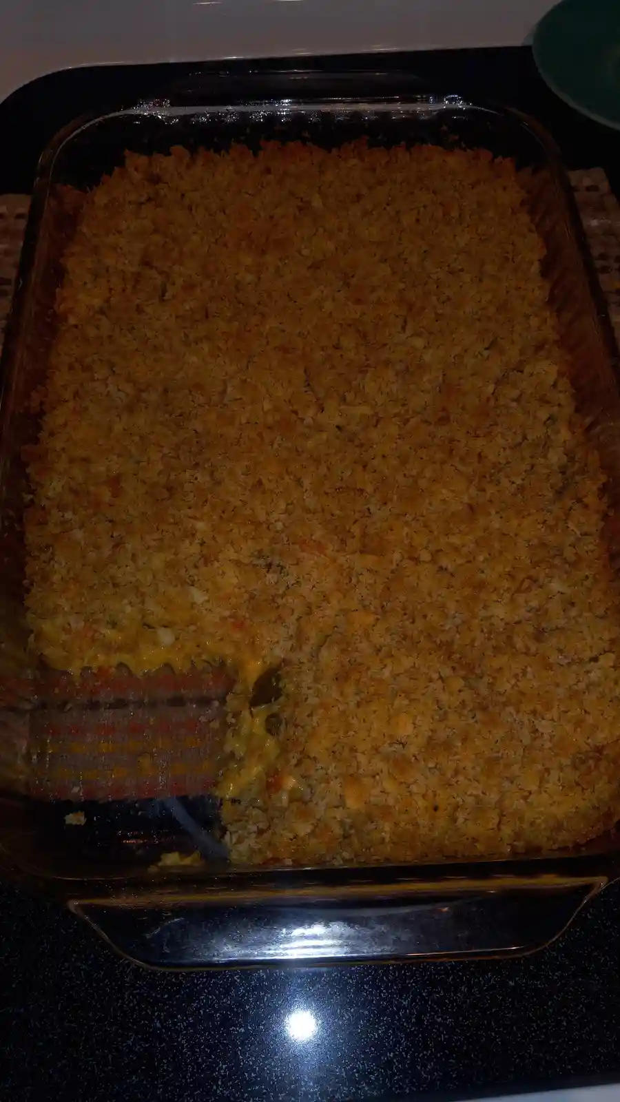 A close-up photo shows a rectangular glass baking dish filled with a golden-brown, crumb-topped casserole. A large portion…