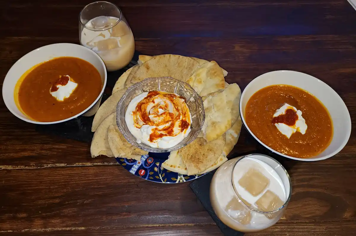 An overhead shot of a meal on a dark wooden table, featuring two bowls of orange-colored curry with dollops of white yogurt…