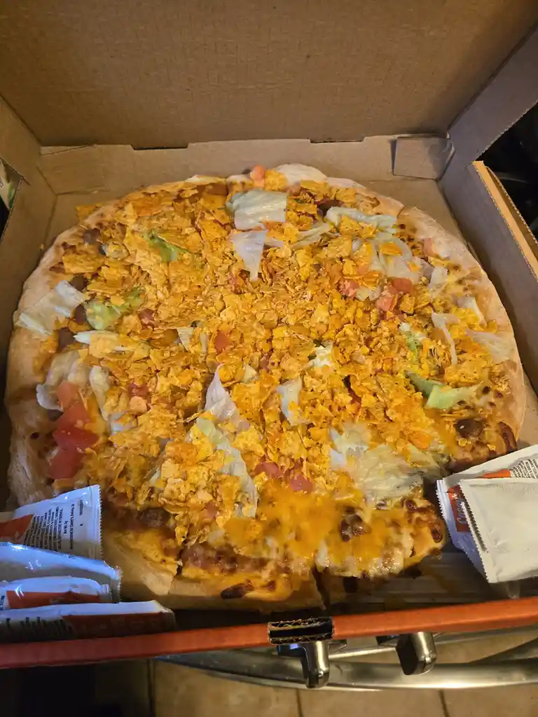 A pizza in an open cardboard box is topped with melted cheese, shredded lettuce, diced tomatoes, and a heavy layer of orange…