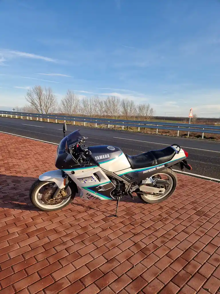 A white, black, and teal Yamaha FZ-1 motorcycle with 'GENESIS SMILE' written on the side is parked on a red brick surface…